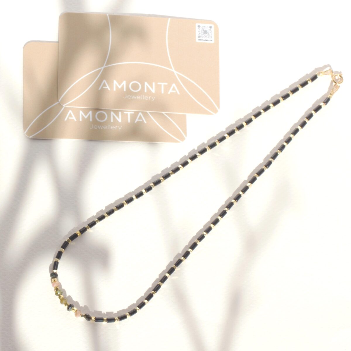 Black Spinel and Pyrite gemstone necklace styled on a white surface with soft shadows, next to AMONTA Jewellery branding cards, showcasing minimalist design and elegant contrast