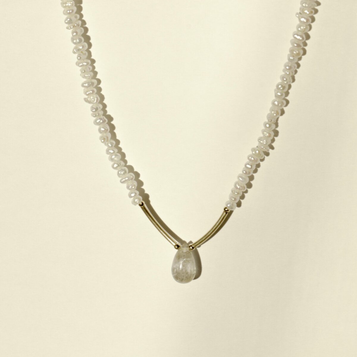 Gold filled Peacock Pearl necklace with a clear teardrop gemstone pendant and curved gold accents, photographed on a warm cream background.