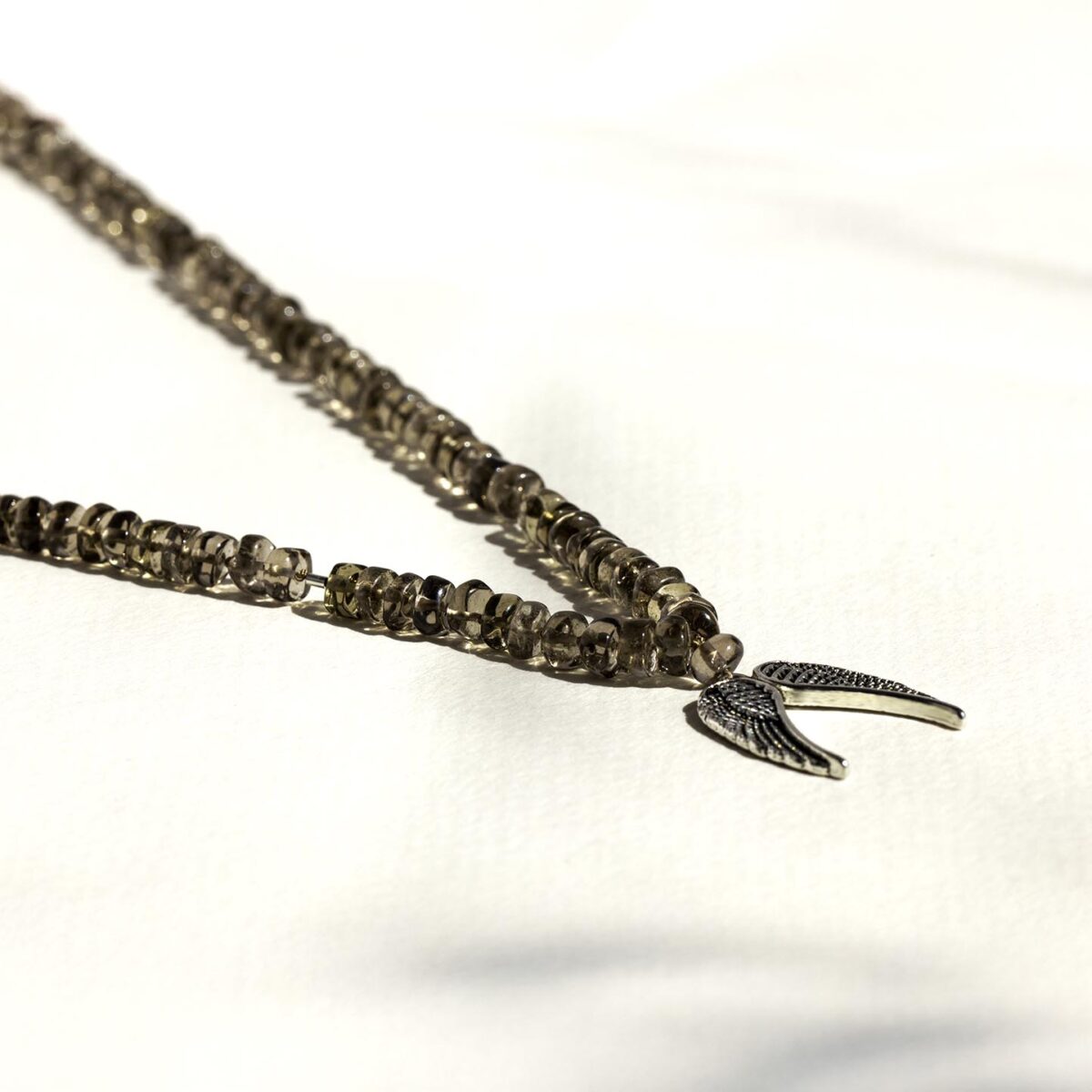 Close-up of silver wing gemstone necklace with smoky quartz beads and detailed sterling silver angel wing pendant, resting on a soft white surface