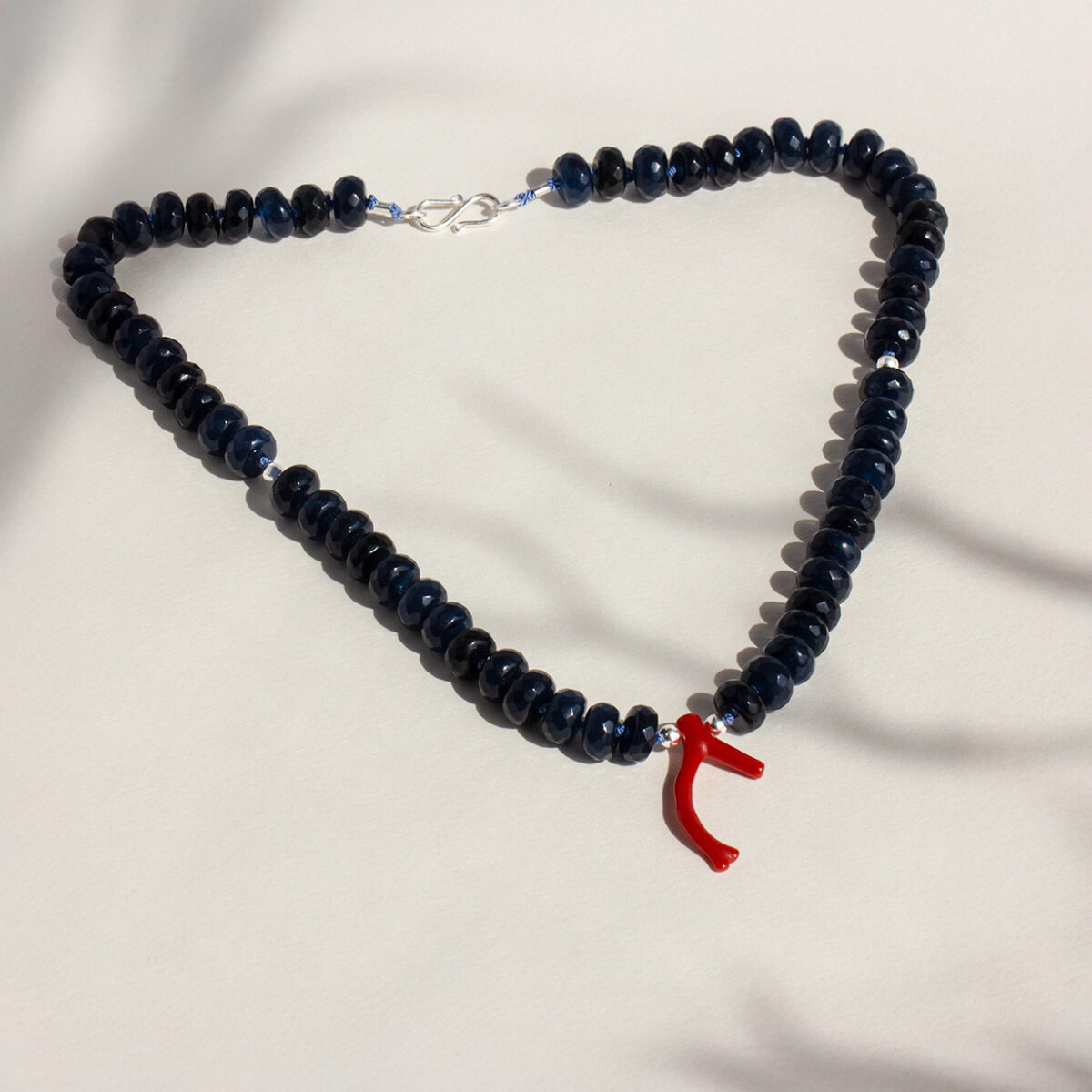 Close-up of the Blue Sea Necklace, featuring smooth round Blue Jade beads and a striking red Coral Branch pendant, hand-strung on silk thread with a silver clasp, displayed against a neutral background with soft shadows.