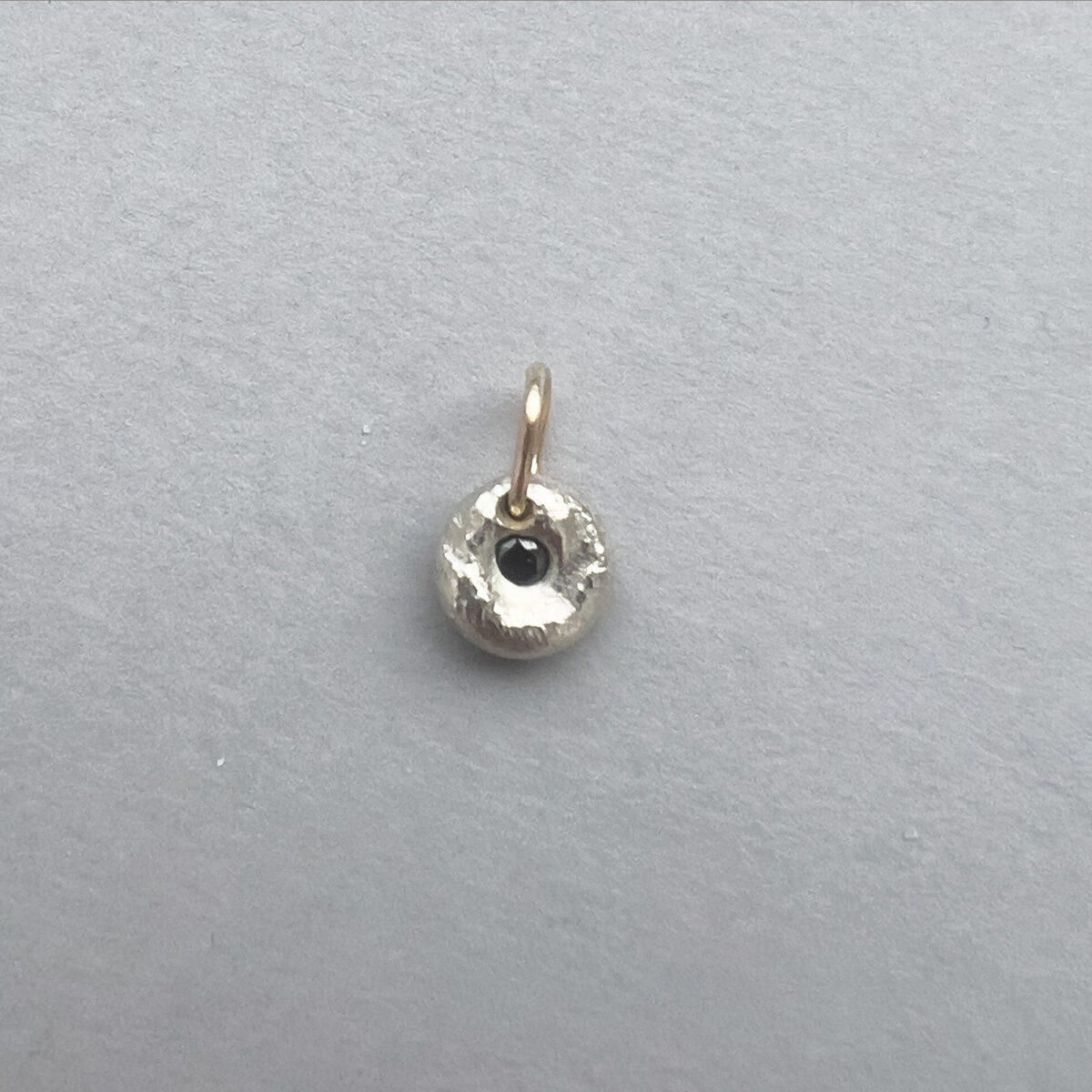 Small round Argentium silver pendant with textured surface, set with a central black diamond and finished with a simple gold bail.