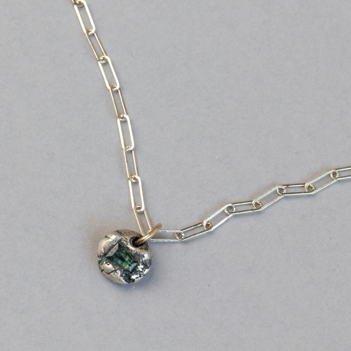 Green rectangular sapphire set in a textured round silver pendant on a fine gold paperclip chain against a grey background.